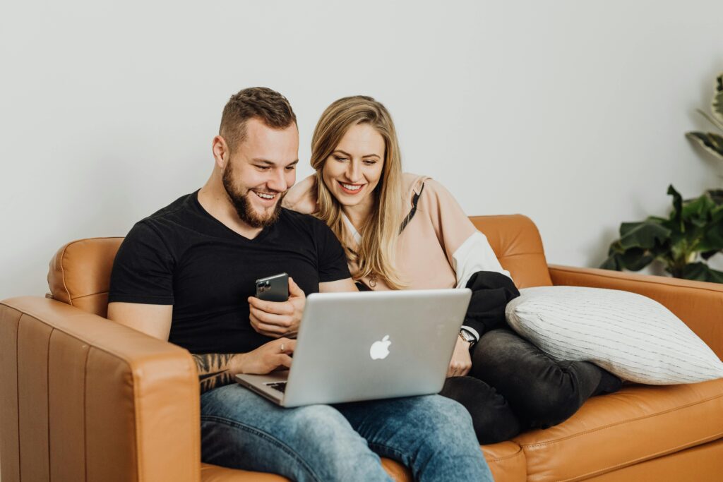 Paar sitzt entspannt am Sofa und bespricht etwas über Laptop und Smartphone.