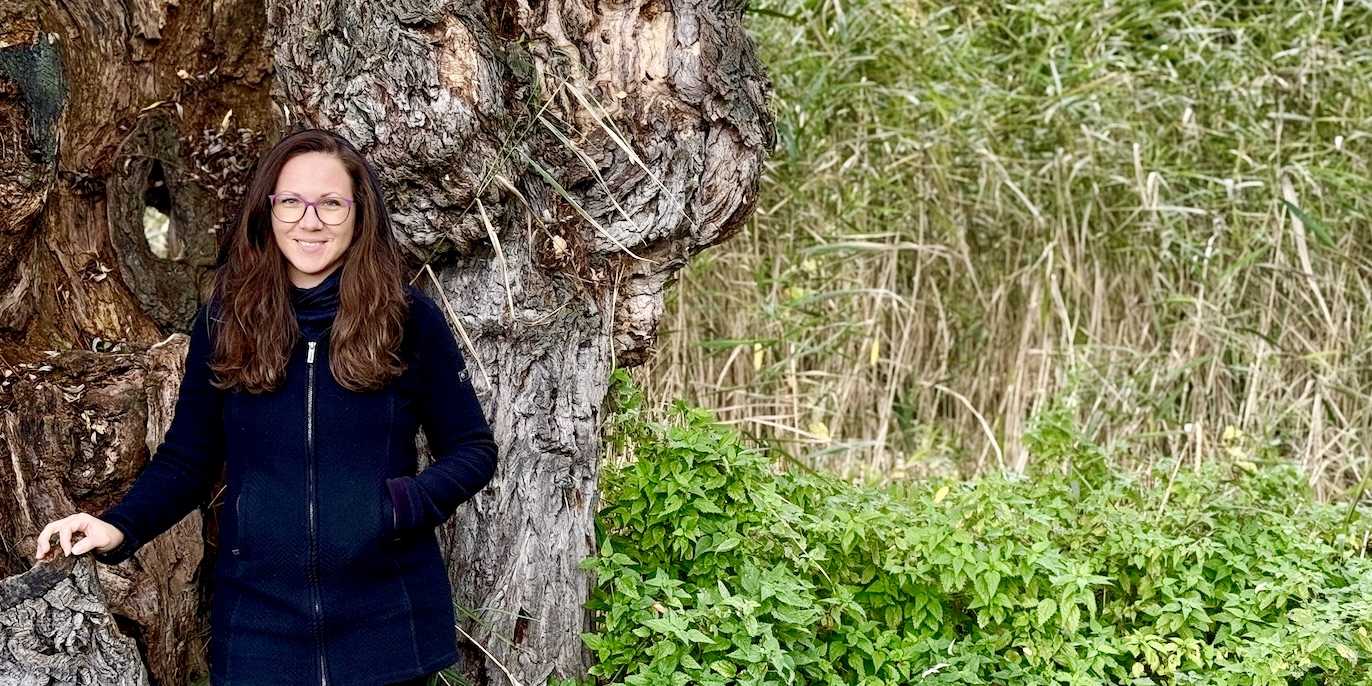 Tanja Hackl steht lächelnd vor einem alten Baumstamm und strahlt Ruhe, Natürlichkeit und Verbundenheit mit der Natur aus.